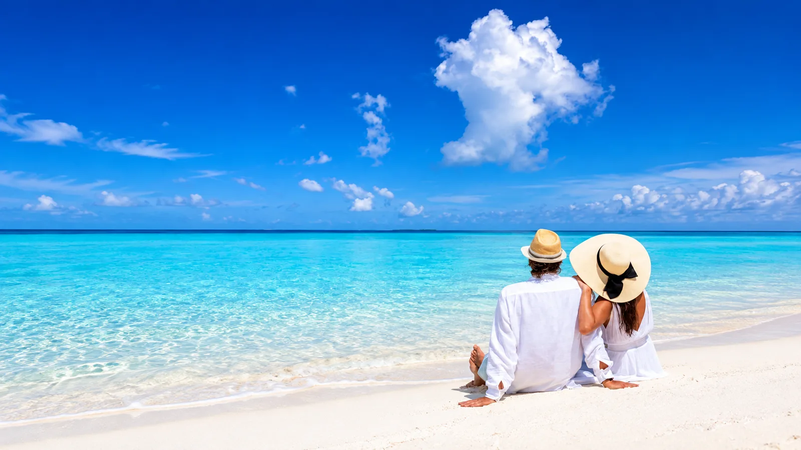 A couple in white sitting together on a white-sand beach, looking out at turquoise water
