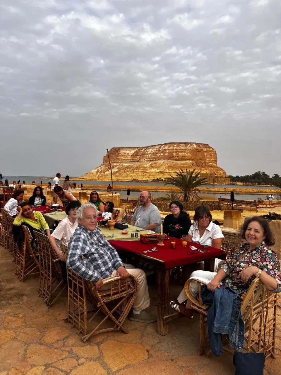 Travellers seated at a long table at sunset, with a flat-topped desert mesa across the water